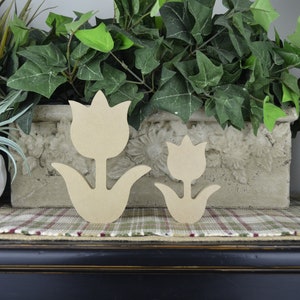 Op de afbeelding: Twee houten tulpenvormen, een groter dan de andere, worden getoond op een geruit oppervlak. De tulpen zijn ongeverfd en hebben een natuurlijke houtkleur.