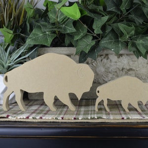 May include: Two wooden cutout buffalo silhouettes, one larger than the other, are displayed on a plaid fabric surface. The larger buffalo is standing with its head turned to the side, while the smaller buffalo is standing with its head facing forward. Both buffalo are painted in a light brown color.