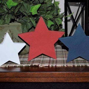 May include: Three wooden stars in red, white, and blue, arranged in a row on a plaid surface. The stars are a patriotic decoration.