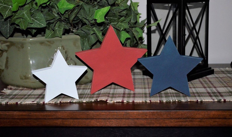 May include: Three wooden stars in red, white, and blue, arranged in a row on a plaid surface. The stars are a patriotic decoration.