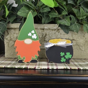 May include: A green leprechaun gnome with an orange beard and a pot of gold. The gnome is wearing a green hat with shamrocks. The pot of gold has a white ribbon and shamrocks.
