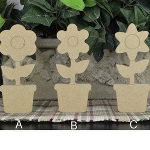May include: Three wooden flower cutouts in pots. Each flower has a different petal shape. The cutouts are beige and sit on a checkered surface. The background features green foliage.