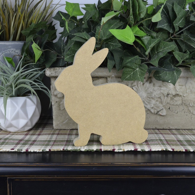 May include: A wooden cutout of a rabbit in a sitting position. The cutout is made of unfinished wood and is a light brown color.