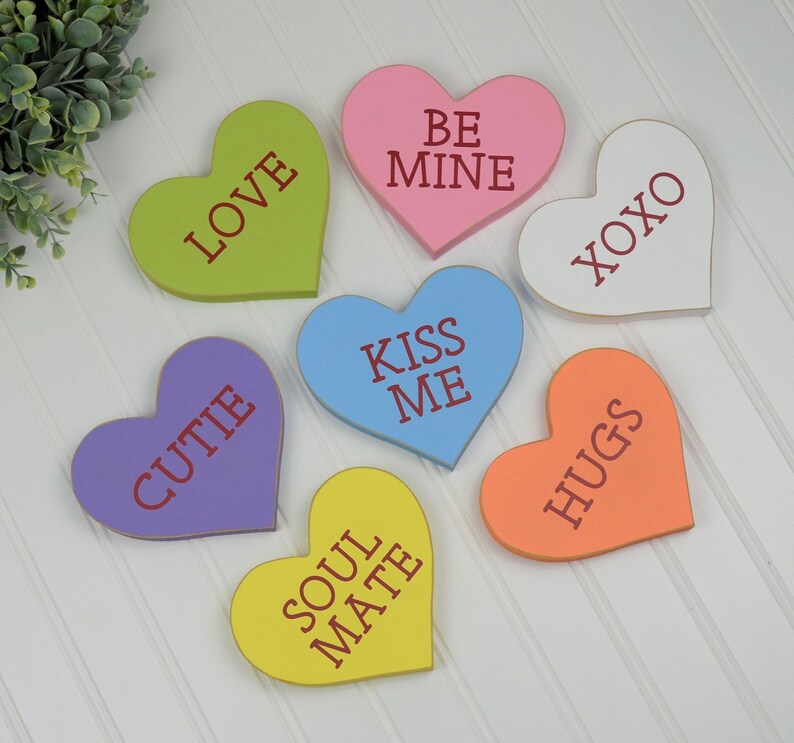 Op de afbeelding: Zes houten hartvormige decoraties met verschillende spreuken op elk. De spreuken zijn "Love", "Be Mine", "XOXO", "Kiss Me", "Cutie" en "Soul Mate".