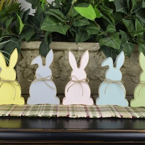 May include: Seven wooden bunny figurines in various pastel colors, including light blue, yellow, gray, pink, light green, and purple. Each bunny has a simple, rustic design and a twine bow around its waist.