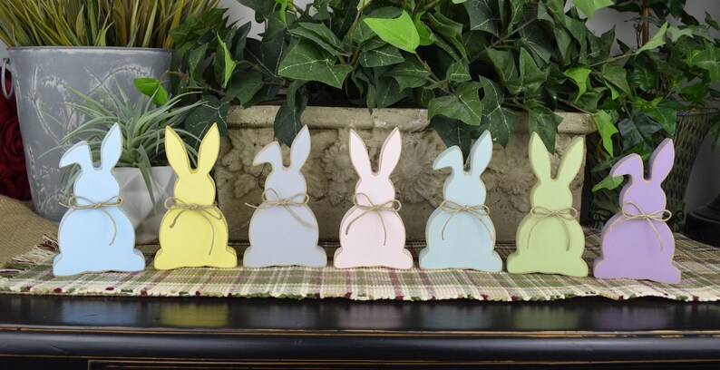 May include: Seven wooden bunny figurines in various pastel colors, including light blue, yellow, gray, pink, light green, and purple. Each bunny has a simple, rustic design and a twine bow around its waist.