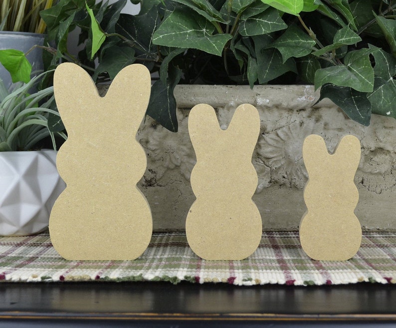 May include: Three unfinished wooden bunny cutouts of varying sizes. The bunnies are light brown and have a simple, classic design. They are standing on a plaid patterned surface, with greenery in the background. Perfect for Easter crafts and decorations.