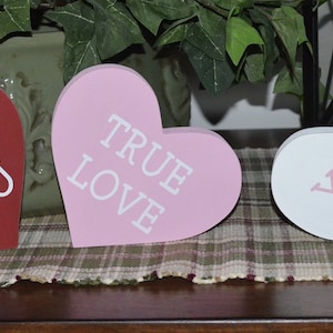 Puede incluir: Tres letreros de madera en forma de coraz&oacute;n con diferentes mensajes. El primer letrero es rojo con texto blanco que dice "I &hearts; YOU". El segundo letrero es rosa con texto blanco que dice "TRUE LOVE". El tercer letrero es blanco con texto rosa que dice "YOU & ME".