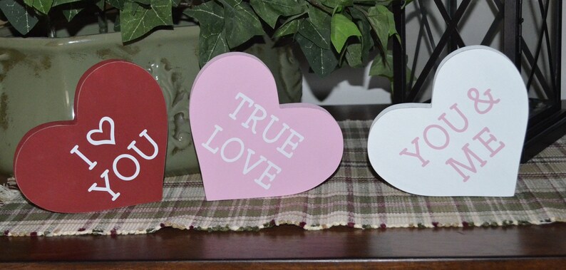 Puede incluir: Tres letreros de madera en forma de coraz&oacute;n con diferentes mensajes. El primer letrero es rojo con texto blanco que dice "I &hearts; YOU". El segundo letrero es rosa con texto blanco que dice "TRUE LOVE". El tercer letrero es blanco con texto rosa que dice "YOU & ME".