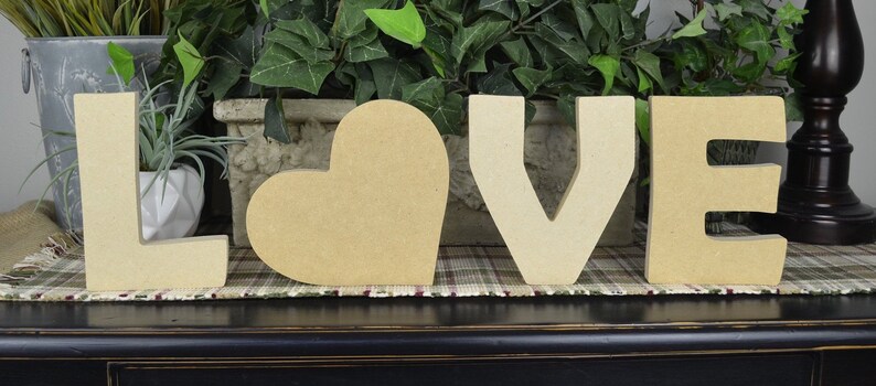 May include: Wooden letters spelling "LOVE" with a heart in the middle. The letters are beige and the heart is beige. The letters are on a plaid surface.