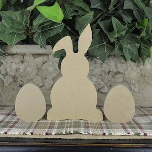 May include: Three unfinished wooden craft shapes: a bunny and two eggs. The bunny is sitting upright with its ears up. The eggs are round and smooth. The shapes are all made of light brown wood.