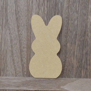May include: A brown wooden cutout of a bunny rabbit in the shape of a Peeps marshmallow candy. The cutout is on a woodgrain background.