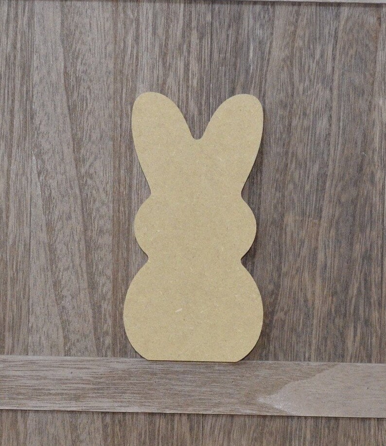 May include: A brown wooden cutout of a bunny rabbit in the shape of a Peeps marshmallow candy. The cutout is on a woodgrain background.