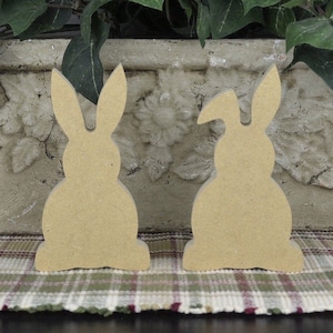 May include: Two unfinished wooden bunny shapes, one with its ears up and the other with one ear down, sitting on a plaid surface.