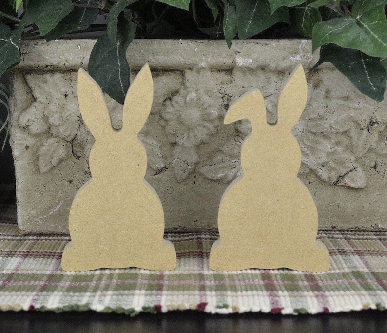 May include: Two unfinished wooden bunny shapes, one with its ears up and the other with one ear down, sitting on a plaid surface.