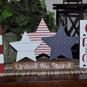 May include: Three wooden stars in red, white, and blue with a wooden sign that reads "United We Stand". The stars are decorated with stripes, polka dots, and a wood grain pattern. The sign is on a brown surface.