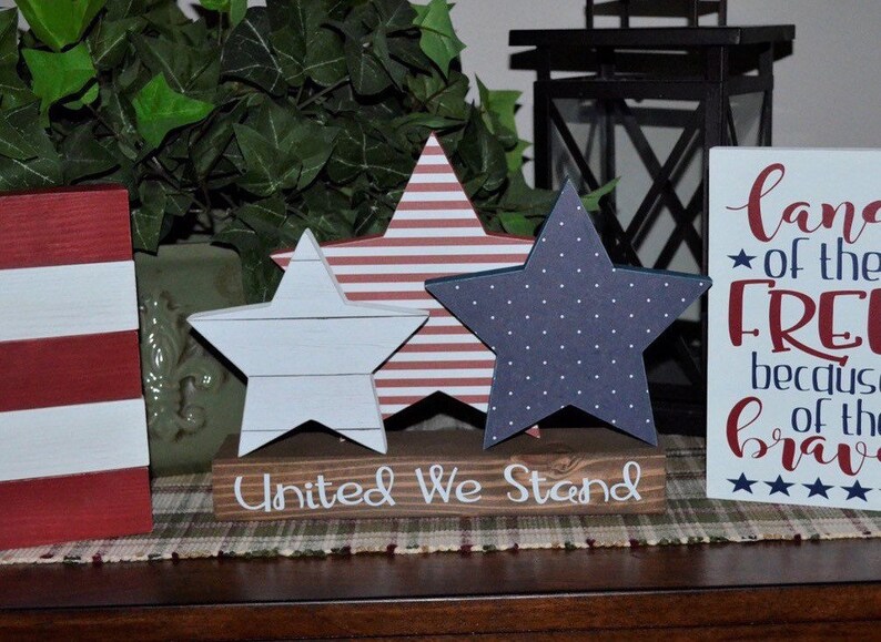 May include: Three wooden stars in red, white, and blue with a wooden sign that reads "United We Stand". The stars are decorated with stripes, polka dots, and a wood grain pattern. The sign is on a brown surface.