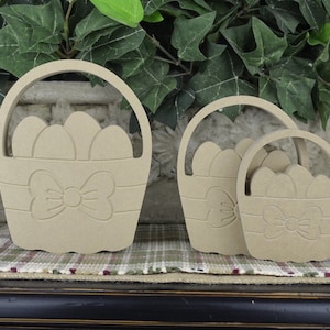 May include: Three unfinished wooden Easter baskets of different sizes, each with a bow and a design of eggs inside. The baskets are arranged from largest to smallest.