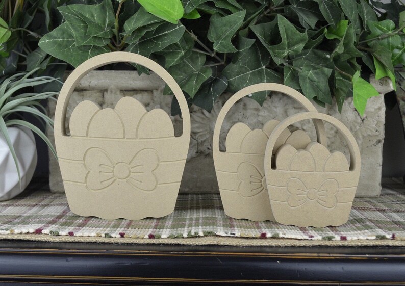 May include: Three unfinished wooden Easter baskets of different sizes, each with a bow and a design of eggs inside. The baskets are arranged from largest to smallest.