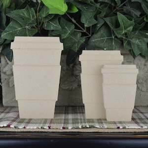 May include: Three unfinished wooden coffee cup shapes in different sizes. The cups are stacked on top of each other and are made of a light brown wood.