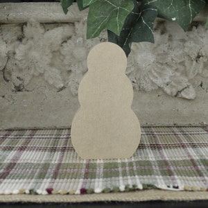 May include: A wooden snowman shape, unpainted, with three rounded sections. The snowman is sitting on a plaid fabric surface.