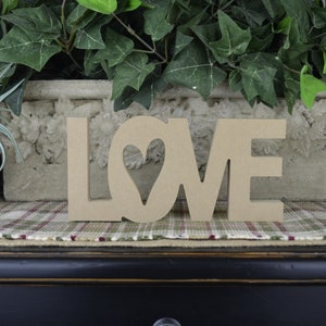 Op de afbeelding: Houten uitgesneden letters die "LOVE" spellen met een hartvormige uitsparing in de "O". De letters liggen op een geruit stofoppervlak.