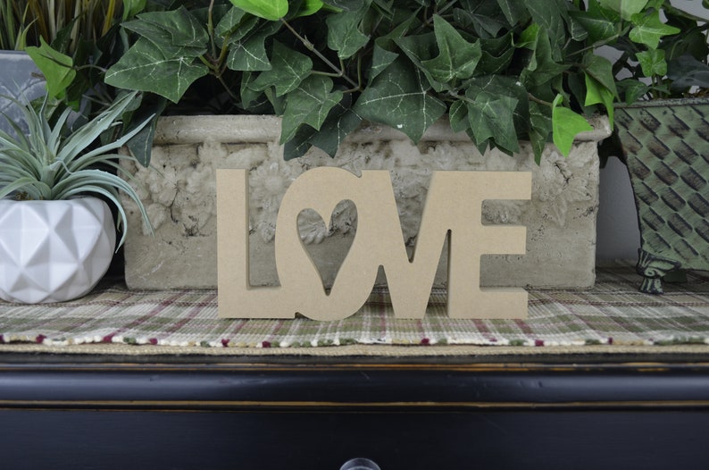 Op de afbeelding: Houten uitgesneden letters die "LOVE" spellen met een hartvormige uitsparing in de "O". De letters liggen op een geruit stofoppervlak.