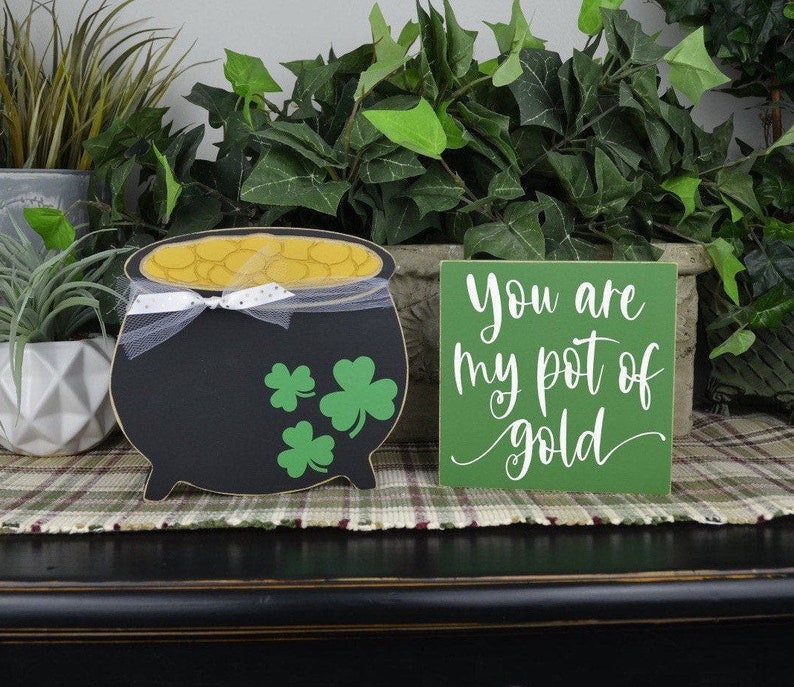 May include: A black pot of gold with green shamrocks and a white ribbon, next to a green sign that reads "You are my pot of gold".