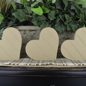 Op de afbeelding: Drie houten harten met een gestreept patroon, gerangschikt in een rij op een geruit oppervlak. De harten zijn lichtbruin van kleur en zijn gemaakt van hout.