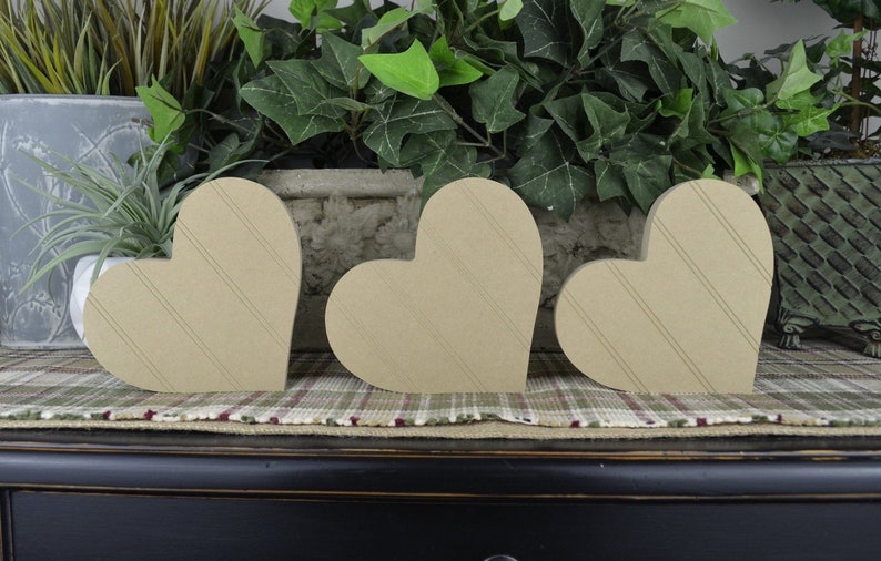 Op de afbeelding: Drie houten harten met een gestreept patroon, gerangschikt in een rij op een geruit oppervlak. De harten zijn lichtbruin van kleur en zijn gemaakt van hout.