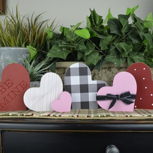 Puede incluir: Cinco decoraciones de madera en forma de coraz&oacute;n en varios colores y patrones. Los corazones est&aacute;n dispuestos en una fila sobre una superficie de madera. Los corazones presentan un patr&oacute;n a cuadros en blanco y negro, un coraz&oacute;n rosa con un lazo negro, un coraz&oacute;n rojo con lunares blancos, un coraz&oacute;n blanco con rayas rosas y un coraz&oacute;n rojo con las palabras "Be Mine" en blanco.