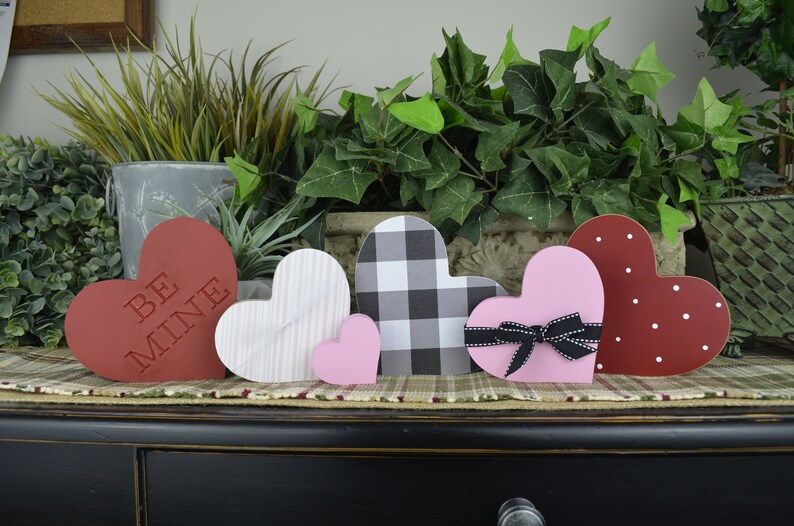 Puede incluir: Cinco decoraciones de madera en forma de coraz&oacute;n en varios colores y patrones. Los corazones est&aacute;n dispuestos en una fila sobre una superficie de madera. Los corazones presentan un patr&oacute;n a cuadros en blanco y negro, un coraz&oacute;n rosa con un lazo negro, un coraz&oacute;n rojo con lunares blancos, un coraz&oacute;n blanco con rayas rosas y un coraz&oacute;n rojo con las palabras "Be Mine" en blanco.