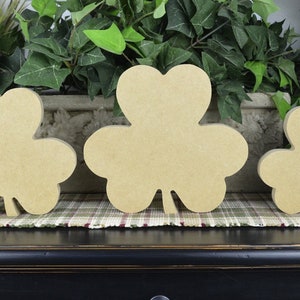 May include: Three unfinished wooden shamrock cutouts of varying sizes. The shamrocks are light brown and sit on a plaid patterned surface. Green plants are in the background.