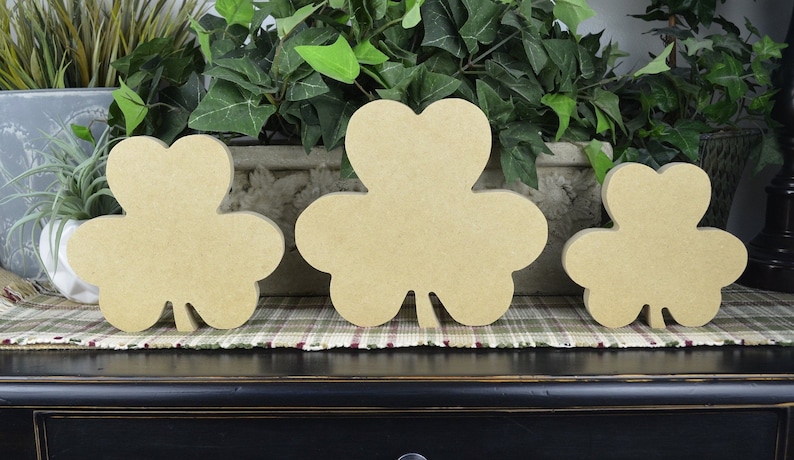 May include: Three unfinished wooden shamrock cutouts of varying sizes. The shamrocks are light brown and sit on a plaid patterned surface. Green plants are in the background.