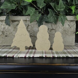 May include: Three unfinished wooden ornaments: a Christmas tree, a large snowman, and a smaller snowman. The ornaments are on a plaid surface.