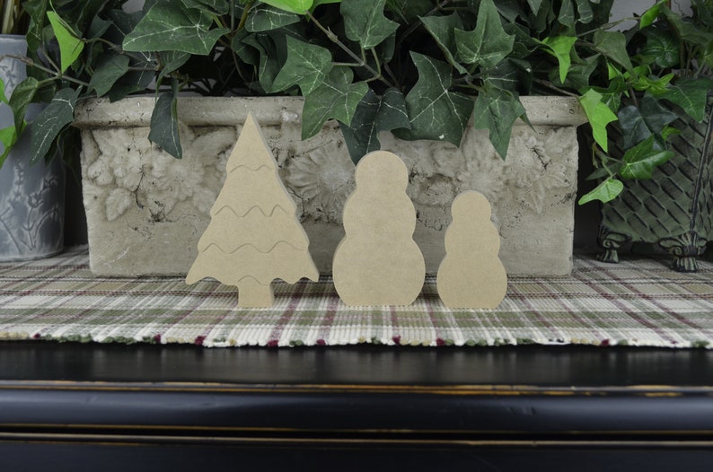 May include: Three unfinished wooden ornaments: a Christmas tree, a large snowman, and a smaller snowman. The ornaments are on a plaid surface.