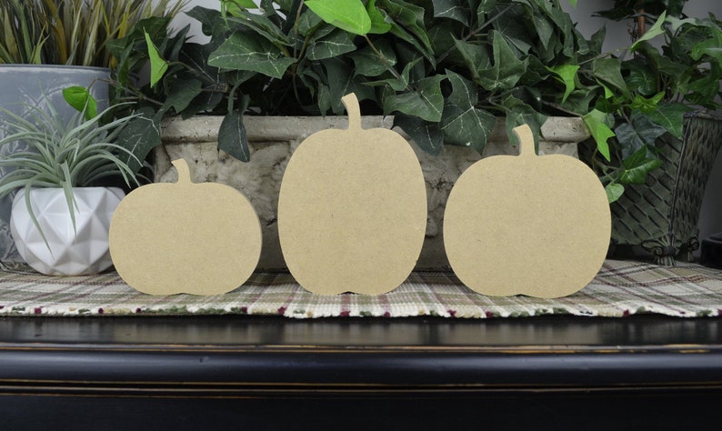 May include: Three unfinished wooden pumpkin shapes of varying sizes. The pumpkins are arranged from largest to smallest, with the largest pumpkin in the center.