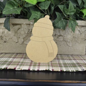 May include: Unpainted wooden snowman cutout with a hat and scarf. The snowman is standing on a plaid surface.