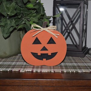 Puede incluir: Calabaza de madera naranja con una cara de calabaza tallada negra. La calabaza tiene un tallo marr&oacute;n y un lazo de rafia.