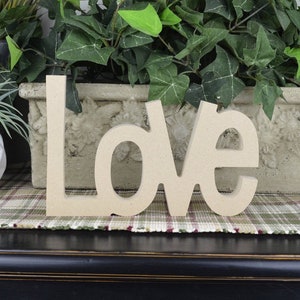 K&ouml;nnte beinhalten: Holzbuchstaben, die "LOVE" in hellbrauner Farbe buchstabieren. Die Buchstaben liegen auf einem karierten Teppich.