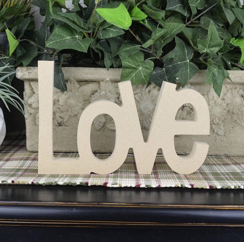 K&ouml;nnte beinhalten: Holzbuchstaben, die "LOVE" in hellbrauner Farbe buchstabieren. Die Buchstaben liegen auf einem karierten Teppich.