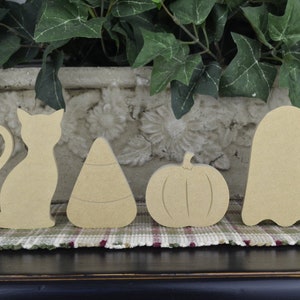 May include: Four unfinished wooden Halloween cutouts: a cat, candy corn, pumpkin, and ghost. The shapes are light brown and sit on a plaid patterned surface. Green foliage and a decorative stone planter are in the background.