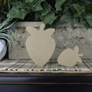May include: Two wooden strawberry shapes, one large and one small, are sitting on a plaid surface. The strawberries are unpainted and have a natural wood color.