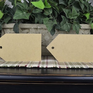 May include: Two unfinished wooden tags with a hole punched in the top. The tags are sitting on a plaid fabric surface.