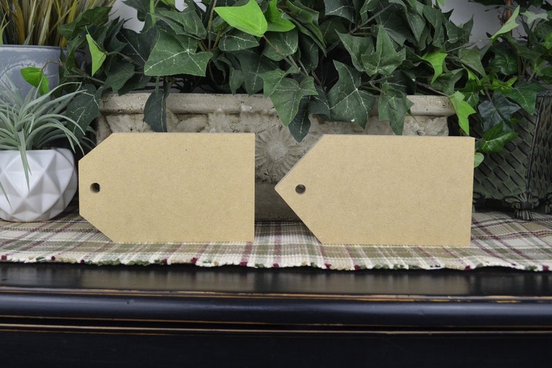 May include: Two unfinished wooden tags with a hole punched in the top. The tags are sitting on a plaid fabric surface.