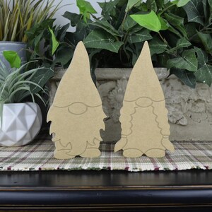 May include: Two unfinished wooden gnome figures, one with a beard and one without, standing on a plaid surface.