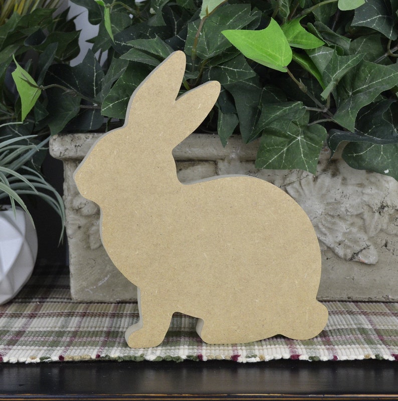 May include: A wooden cutout of a rabbit in a sitting position. The cutout is unpainted and has a natural wood color.