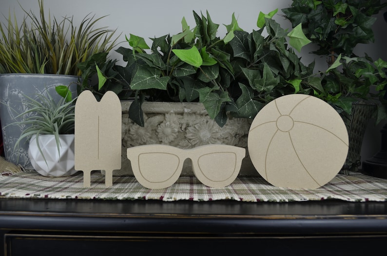 May include: Three unfinished wooden cutouts of summer-themed items: a popsicle, sunglasses, and a beach ball. The items are arranged in a row on a brown and white checkered surface.