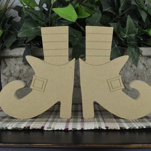 May include: Two wooden cutouts of elf shoes, each with a curled toe, a buckle detail, and a tall, striped boot. The shoes are a light tan color and are displayed on a plaid patterned surface.
