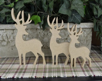 May include: Three wooden deer silhouettes of varying sizes. The deer are light brown and have detailed antlers. They stand on a plaid patterned surface in front of a decorative planter with green foliage.